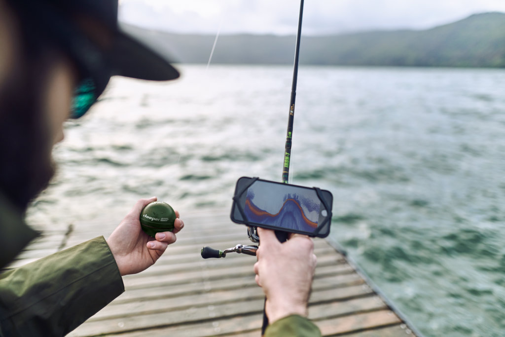 Angler mit einem Deeper-Fischfinder und einer Angel in der Hand steht auf einem Steg.