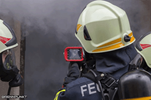 Two firefighters are standing in front of a house entrance from which dense smoke is coming. One of the two firefighters looks into the building entrance through his thermal imaging camera.