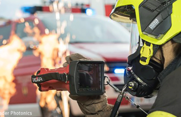Ein Feuerwerhmann benutzt eine Wärmebildkamera. Im Hintergrund ist ein Feuerwehrauto zu sehen und es sprühen Funken