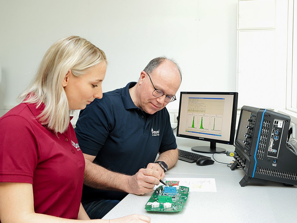 Produktmanagerin Leonie Weißer und Ingenieur Mathias Laskus schauen sich eine Leiterplatte genau an. Im Hintergrund sind ein Bildschirm und ein Oszilloskop zu sehen.