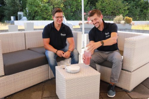 Zwei Männer sitzen gemeinsam auf einer Terrasse und mixen einen Smoothie
